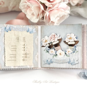 May include: A light blue and white scrapbook with a sewing theme. The front cover features a sewing pin holder with a banner that says "Sewing Pins". The back cover features a floral design with spools of thread and a banner that says "Thread".