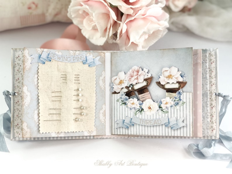 May include: A light blue and white scrapbook with a sewing theme. The front cover features a sewing pin holder with a banner that says "Sewing Pins". The back cover features a floral design with spools of thread and a banner that says "Thread".
