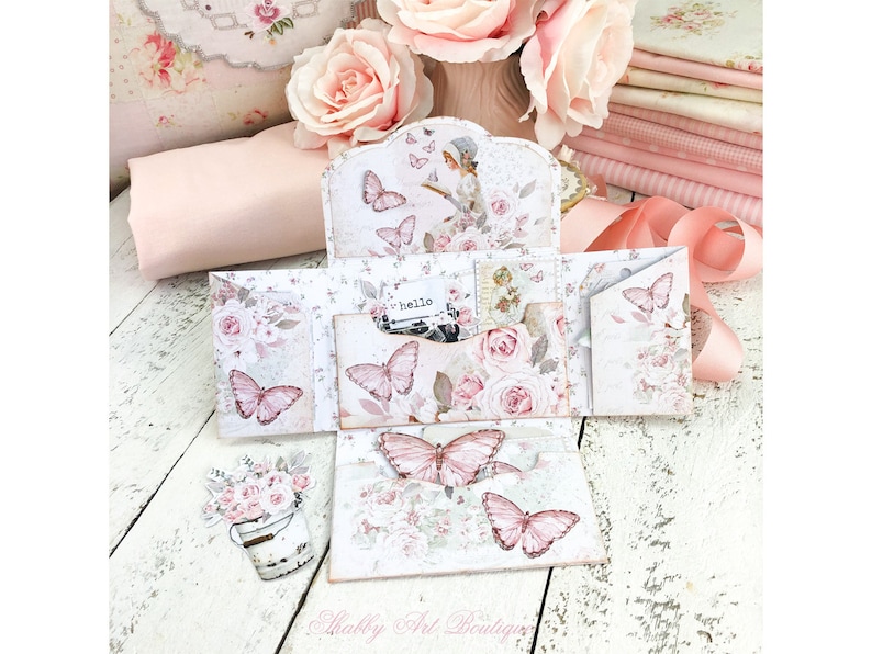 May include: A white and pink paper craft with a floral design and butterflies. The craft is folded into a series of panels and has a small pocket on the front. The text "hello" is visible on a small card inside the pocket.