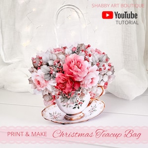 May include: A decorative teacup bag filled with pink and red flowers and berries. The teacup and saucer are white with gold trim and floral designs. The bag has a handle and the text "Christmas Teacup Bag" is visible.
