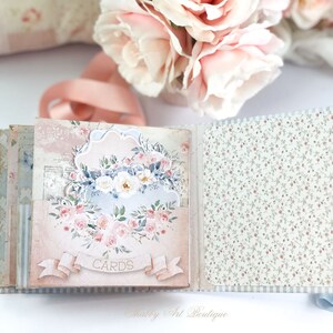 May include: A pink and blue scrapbook with a floral design and the word "CARDS" on the front. The scrapbook is open to reveal a page with a floral pattern.