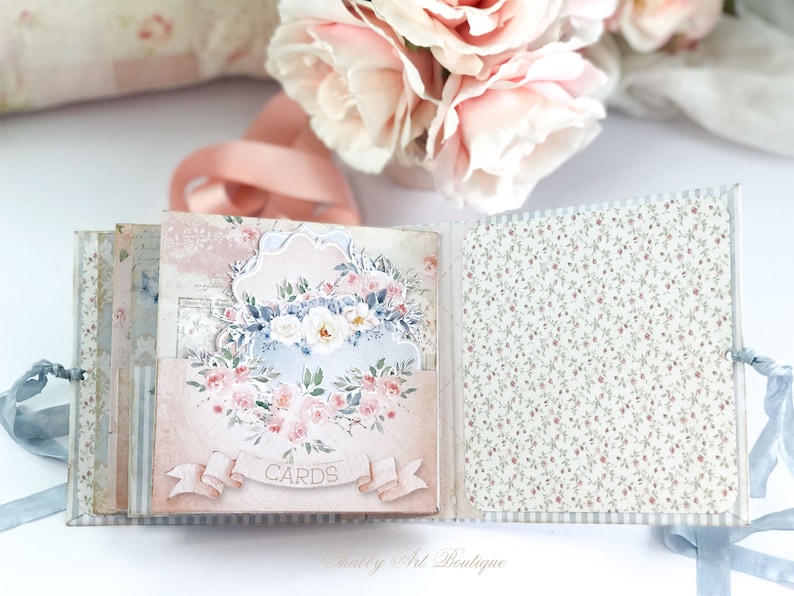 May include: A pink and blue scrapbook with a floral design and the word "CARDS" on the front. The scrapbook is open to reveal a page with a floral pattern.