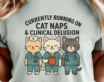 May include: A light green t-shirt featuring the text "Currently Running on Cat Naps & Clinical Delusion." The shirt has a graphic of three cartoon cats dressed as medical professionals. The cats are wearing scrubs and holding a coffee mug.