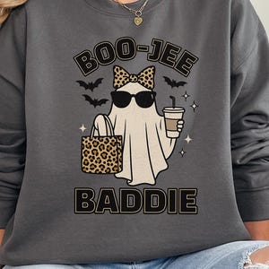 May include: Charcoal gray sweatshirt with a graphic of a ghost wearing sunglasses and a bow, holding a leopard print bag and a coffee cup. The text "BOO-JEE BADDIE" is above and below the ghost. The sweatshirt is a Gildan brand.