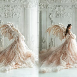 May include: A pregnant person wearing a flowing, blush-coloured tulle gown with large, feathery angel wings. The gown has a long train and the person is standing in front of a white, ornate wall.
