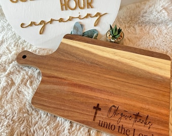 Puede incluir: Una tabla de cortar de madera con asa, grabada con el texto "Oh give thanks unto the Lord..." y una cruz.