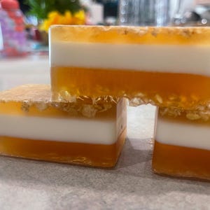 May include: Three layered bars of soap with a white, orange, and yellow color scheme. The soap has a textured surface and is made with natural ingredients.