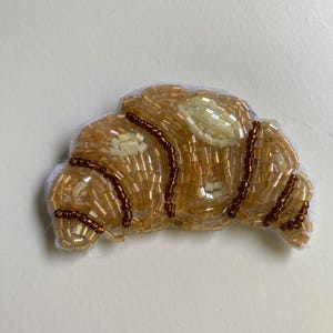 May include: A beaded croissant brooch featuring brown and gold beads. The croissant is crafted from light brown beads with white beads representing the flaky layers.