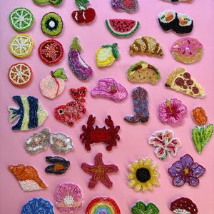 May include: A selection of beaded patches in diverse forms, such as fruits, sushi, flowers, and marine life. The patches display vivid colours and detailed beadwork, set against a pink backdrop.