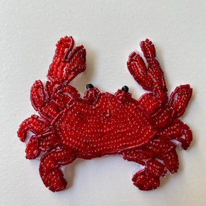 May include: A vibrant red beaded crab applique with black eyes and a white backing. The crab features two prominent claws and is crafted from small, glossy beads. Ideal for adding a seaside theme to garments or accessories.