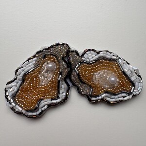 May include: Two geode-shaped brooches, intricately beaded. The centres are filled with gold and bronze beads, each with a pearl. The edges are outlined with white, silver, and black beads. The brooches are set against a white backdrop.