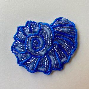 May include: A handcrafted, blue beaded seashell brooch. The brooch features a spiral design with varying shades of blue and silver beads. The edges are outlined with a darker blue, creating a textured, three-dimensional effect. This accessory is perfect for adding a touch of coastal charm.
