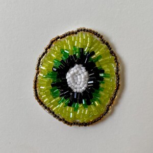 May include: A beaded kiwi fruit slice. The centre is white, surrounded by black, green, and light green beads. The outer edge is a ring of brown beads, and the outer flesh is light green. The beaded fruit is on a white background.
