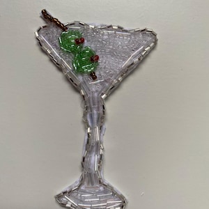 May include: A beaded martini glass with two green olives and a brown stem. The glass is made of white beads and the olives are green with red centres.