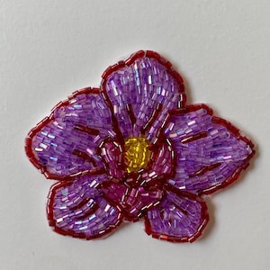 May include: A beaded flower applique with a vibrant design. The petals are a gradient of purple, with a red outline and a yellow centre. The beads create a textured, shimmering effect.