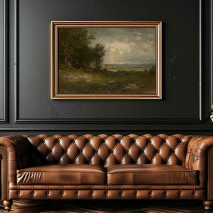 An oil painting in a gold frame depicts a landscape with trees, a field, and a cloudy sky. The painting hangs above a brown leather Chesterfield sofa. The wall color is black.