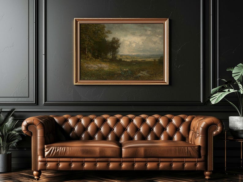An oil painting in a gold frame depicts a landscape with trees, a field, and a cloudy sky. The painting hangs above a brown leather Chesterfield sofa. The wall color is black.