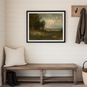 A framed landscape painting with a black frame hangs on a white wall in a modern farmhouse interior entryway. Below the painting is a wooden bench with a cream-colored pillow and black boots. A coat hangs on a wooden coat rack.