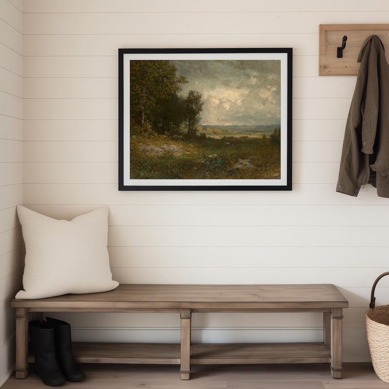 A framed landscape painting with a black frame hangs on a white wall in a modern farmhouse interior entryway. Below the painting is a wooden bench with a cream-colored pillow and black boots. A coat hangs on a wooden coat rack.