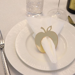 May include: A table setting with white plates, silverware, and a gold apple-shaped napkin ring. The napkin ring holds a white napkin. A wine bottle and glass are in the background. The forks and spoons have a textured design.