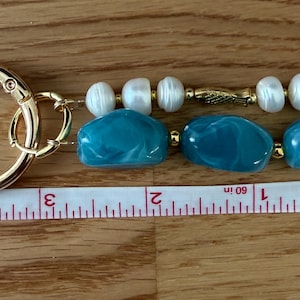 Puede incluir: Un llavero dorado con un cierre redondo grande. El llavero presenta tres cuentas de color turquesa en forma ovalada y tres perlas blancas. El llavero mide aproximadamente 7,6 cm de largo.