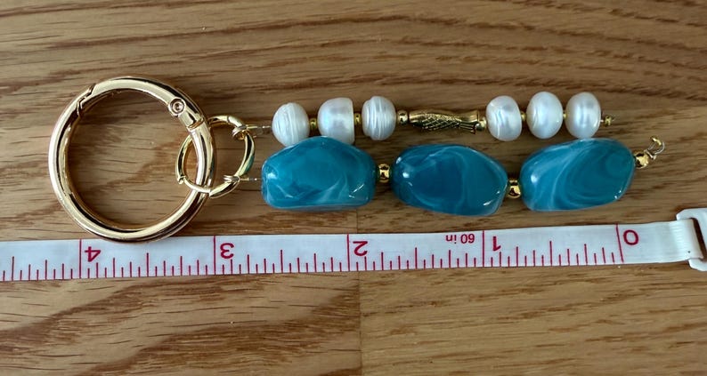Puede incluir: Un llavero dorado con un cierre redondo grande. El llavero presenta tres cuentas de color turquesa en forma ovalada y tres perlas blancas. El llavero mide aproximadamente 7,6 cm de largo.