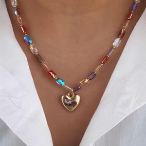 May include: A beaded necklace with a gold heart pendant. The necklace features a variety of colorful, iridescent beads in shades of red, blue, purple, and gold. The heart pendant is smooth and reflective, and the necklace is worn against a white shirt.