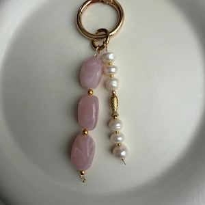 Puede incluir: Un llavero dorado con un cierre redondo. El llavero presenta tres piedras rosas oblongas y una hebra de perlas blancas. Las piedras y las perlas est&aacute;n conectadas con detalles dorados. El llavero est&aacute; sobre un plato blanco.