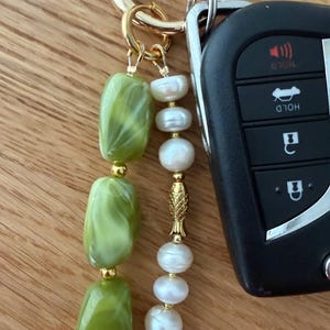 Puede incluir: Un llavero dorado con una cadena de cuentas verdes y blancas veteadas y una cadena de perlas blancas. Un dije de pez dorado separa las perlas. Una llave de coche est&aacute; unida al llavero.