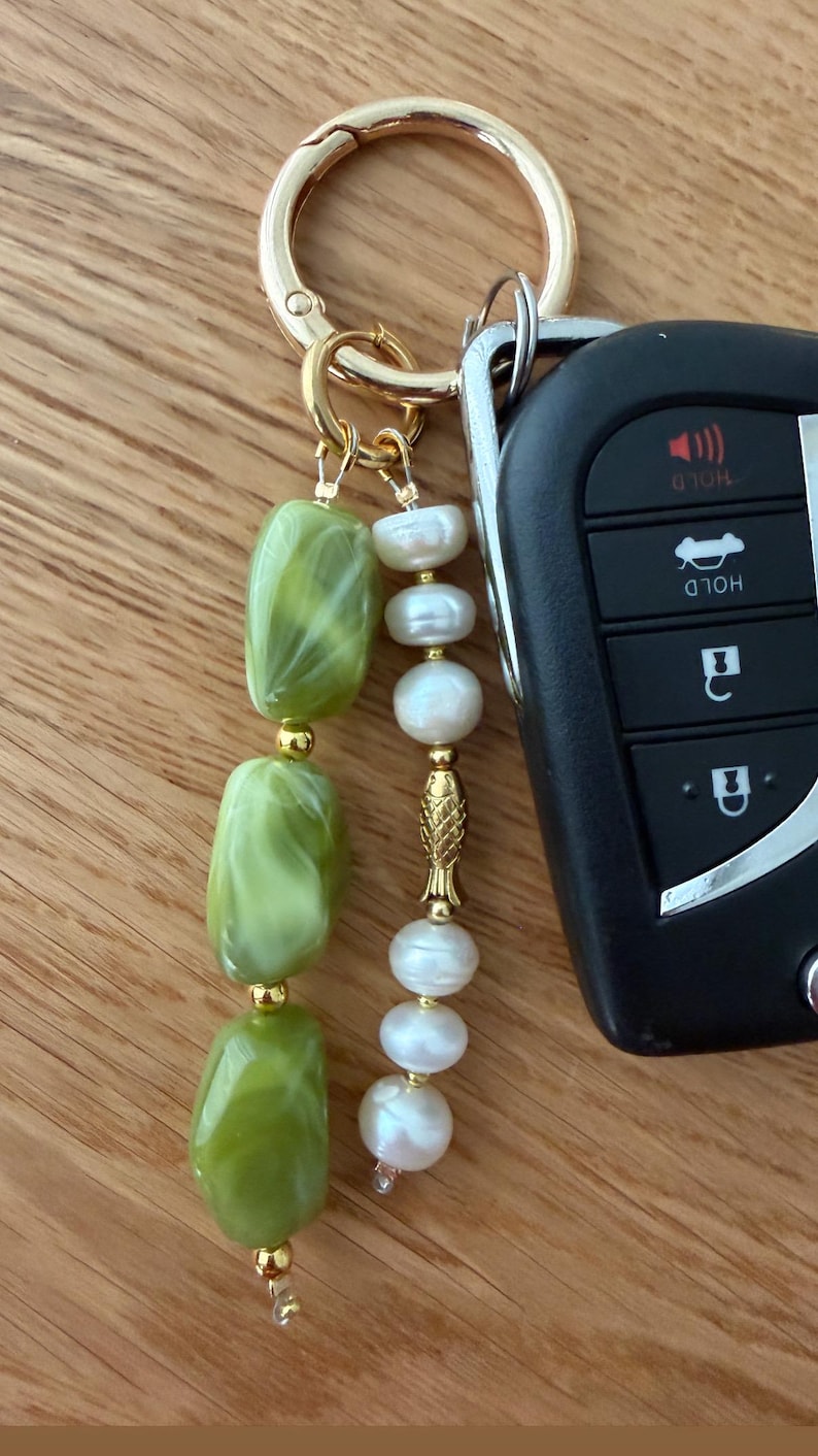 Puede incluir: Un llavero dorado con una cadena de cuentas verdes y blancas veteadas y una cadena de perlas blancas. Un dije de pez dorado separa las perlas. Una llave de coche est&aacute; unida al llavero.