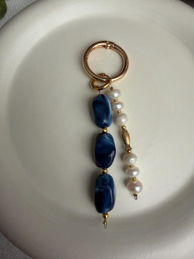 Puede incluir: Un llavero dorado con un cierre grande. El llavero presenta tres cuentas ovaladas de color azul oscuro con vetas blancas y una hebra de perlas blancas. Las cuentas est&aacute;n separadas por peque&ntilde;as cuentas doradas.