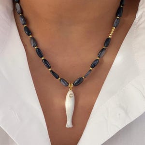 May include: A necklace featuring a pendant shaped like a fish. The pendant is white with a small, round, clear gem. The necklace has dark gray oval beads with gold-colored spacers. The necklace is worn over a white shirt.