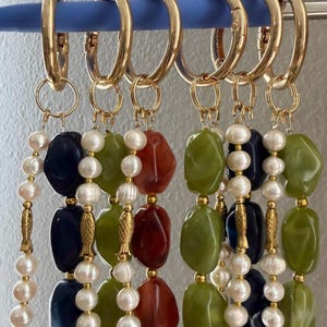 Puede incluir: Pendientes de aro dorados con colgantes de cuentas. Cada colgante presenta una combinaci&oacute;n de piedras de colores de forma irregular, perlas de agua dulce y detalles dorados. Las piedras son en tonos verdes, rojos y azul marino.