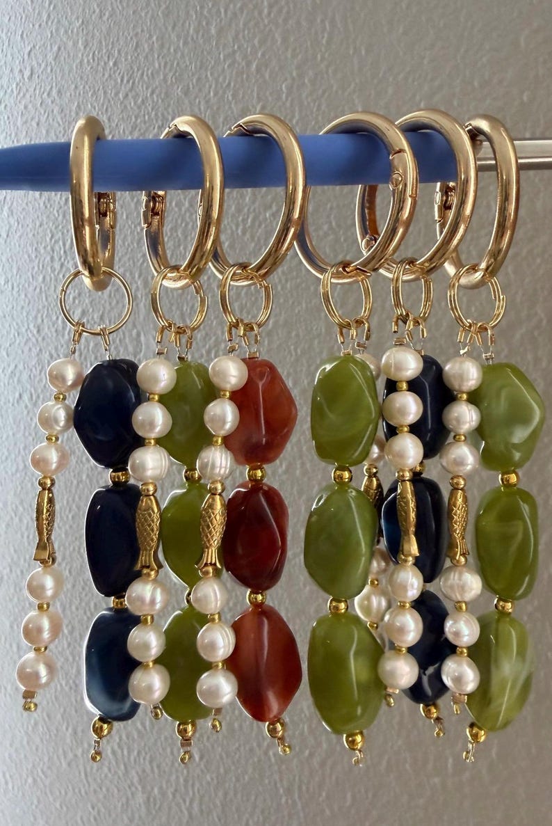 Puede incluir: Pendientes de aro dorados con colgantes de cuentas. Cada colgante presenta una combinaci&oacute;n de piedras de colores de forma irregular, perlas de agua dulce y detalles dorados. Las piedras son en tonos verdes, rojos y azul marino.