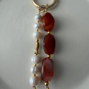Puede incluir: Un llavero dorado con un cierre redondo. El llavero presenta una hebra de perlas blancas y una hebra de piedras oblongas de color marr&oacute;n rojizo. Tambi&eacute;n hay un peque&ntilde;o dije de pez dorado.