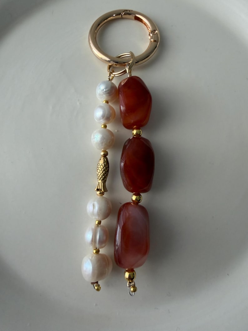 Puede incluir: Un llavero dorado con un cierre redondo. El llavero presenta una hebra de perlas blancas y una hebra de piedras oblongas de color marr&oacute;n rojizo. Tambi&eacute;n hay un peque&ntilde;o dije de pez dorado.