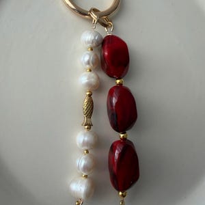 Puede incluir: Un llavero dorado con un cierre de mosquet&oacute;n. El llavero presenta una hebra de perlas blancas y una hebra de tres cuentas rojas oblongas. Un dije de pez dorado y peque&ntilde;as cuentas doradas acent&uacute;an el dise&ntilde;o. El fondo es una superficie blanca.