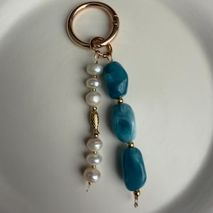 Puede incluir: Un llavero dorado con un cierre redondo. El llavero presenta dos hebras colgantes: una con perlas blancas y un dije de pez dorado, y otra con tres cuentas azul-verdosas de forma irregular. Las cuentas est&aacute;n separadas por detalles dorados.