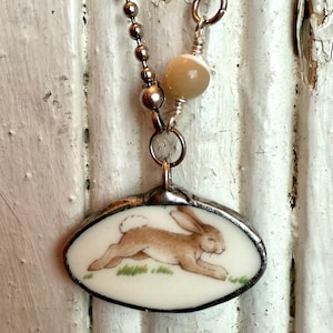 May include: Oval pendant necklace with a running brown rabbit illustration on a white background. The pendant is framed in silver and hangs from a silver ball chain. Beige beads and silver accents complete the necklace.