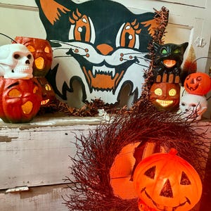 May include: A collection of vintage Halloween decorations, including a large black cat cutout with orange accents, illuminated jack-o'-lanterns, and skull-shaped lights. A dark wreath and a stack of cat and pumpkin figures complete the festive display.
