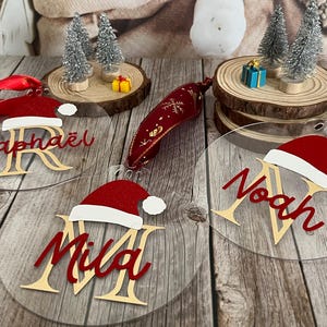 Könnte beinhalten: Klare Weihnachtsornamente aus Acryl mit roten und weißen Weihnachtsmützen, goldenen Buchstabeninitialen und Namen in roter Schrift. Miniatur-Weihnachtsbäume und Geschenke auf Holzscheiben.