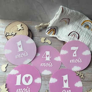 Op de afbeelding: Roze baby mijlpaal schijven met witte cijfers en de woorden "mois" en "an". Elke schijf heeft een hart- en wolkenontwerp. Een witte tas met regenboogpatronen is zichtbaar op de achtergrond.
