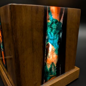 Peut inclure: Un coffret en bois artisanal avec une incrustation de résine vibrante. La résine présente un mélange de teintes orange, turquoise et bleu, contrastant avec le bois foncé. Le coffret repose sur un socle en bois assorti.