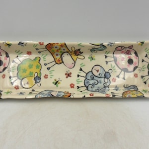 May include: A rectangular ceramic serving dish with a white background and a colourful pattern of cartoon sheep, cows, and butterflies.