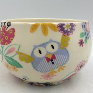 May include: A white ceramic bowl featuring a colourful owl design. The owl is wearing a bow tie and is surrounded by flowers and leaves. The bowl is ideal for serving snacks or desserts.