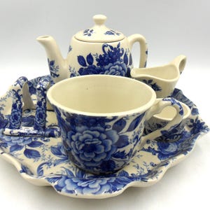 Könnte beinhalten: Ein Vintage-Teeservice mit blauem Blumenmuster. Das Set beinhaltet eine Teekanne, einen Sahnekännchen, eine Tasse und ein Serviertablett. Die Tasse und die Teekanne haben einen weißen Hintergrund mit blauen Blumendesigns. Das Tablett hat einen gewellten Rand.