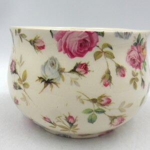 May include: A small, round, cream-coloured ceramic bowl adorned with pink and white roses and green foliage. The floral design covers the entire surface of the bowl, which has a gently curved rim. The bowl is likely for decorative or serving purposes.