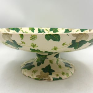 May include: A cream-coloured ceramic pedestal bowl featuring a repeating pattern of green ivy leaves and small clusters of light green berries. The bowl has a wide, shallow basin and a sturdy, flared base, both decorated with the same botanical motif.
