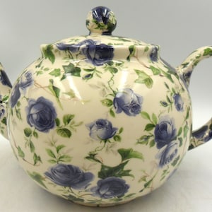 May include: Cream-coloured ceramic teapot with a rounded body, decorated with blue rose floral patterns and green leaves. The teapot has a curved spout, a loop handle, and a lid with a small knob. A decorative item for tea parties.