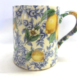 May include: A white ceramic mug with a floral pattern featuring blue flowers, green leaves, and yellow lemons. The mug has a handle.